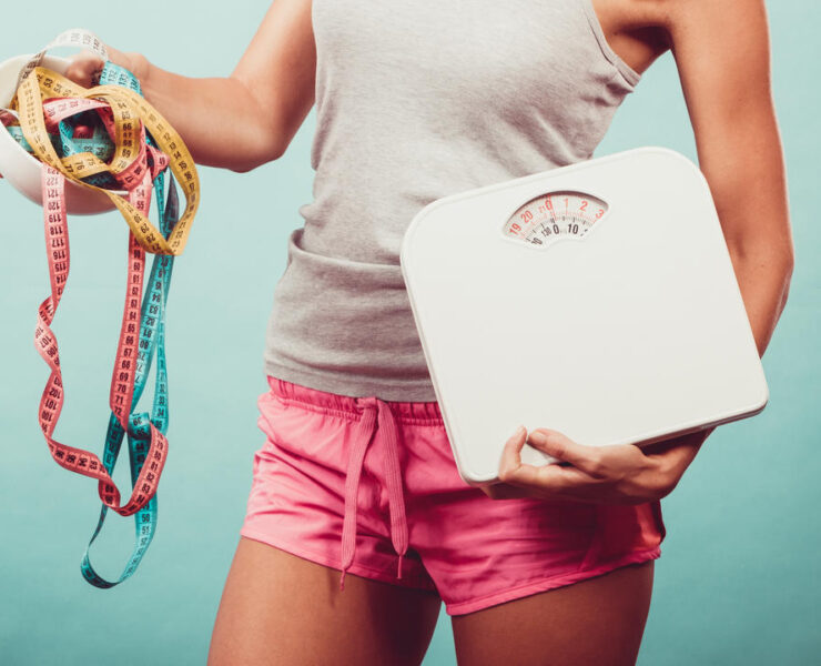 Diet, healthy eating and slim body concept. Fit fitness girl holding bowl with many colorful measuring tapes as dieting symbol and weight scales studio shot on blue